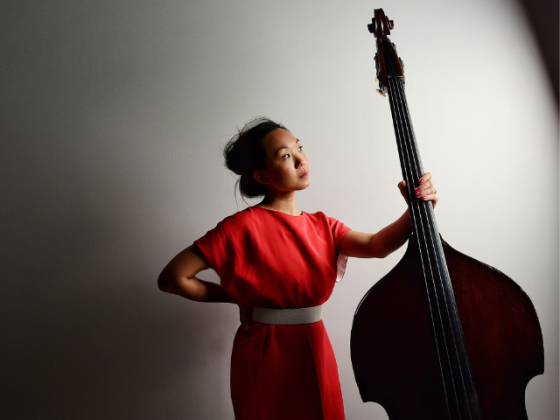 woman with dark hair in a bun and wearing a red dress is holding a double bass by the neck and looking off to the right