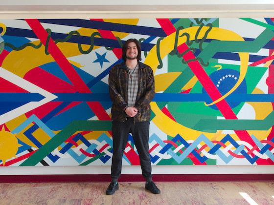 A student standing in front of a colorful, abstract mural.