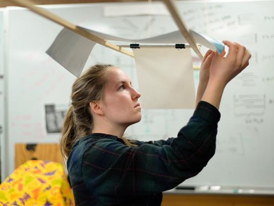 A student inspects a sheet of paper that is draped over 2 wooden rods above her.