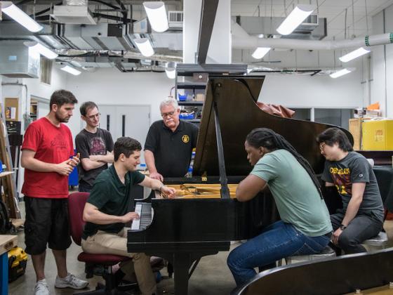 A group of students gather around a piano in a bright workshop.