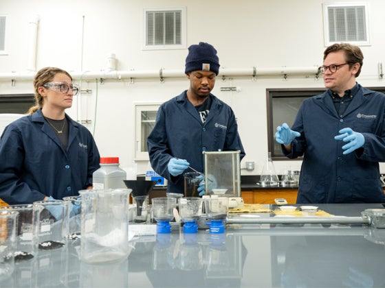 Two students wearing protective gear work in a lab with an employee