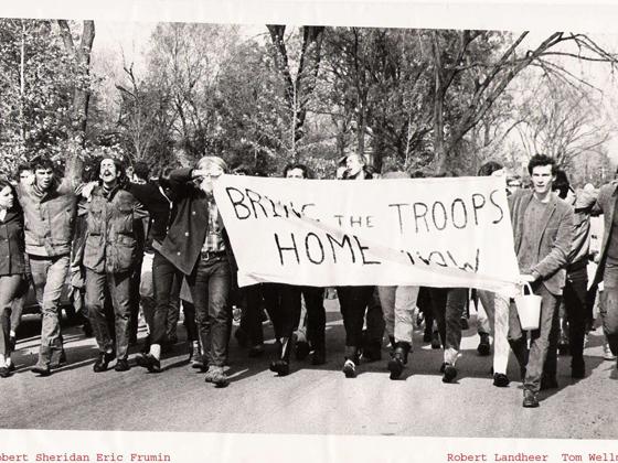 1960s era protesters walk arm in arm, carrying a banner reading 'Bring our troops home now.'