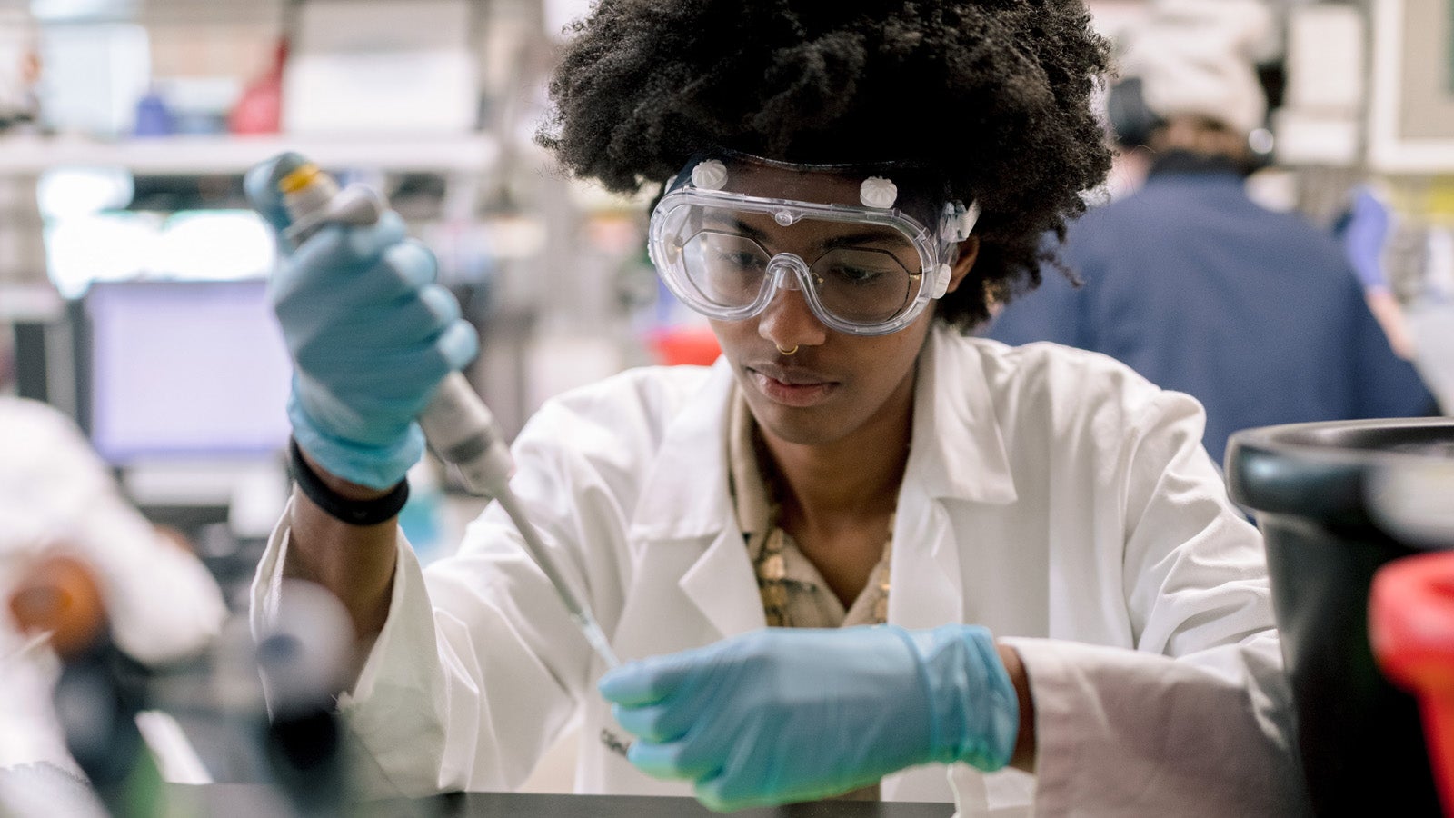 a student wearing a lab coat and blue gloves works in a lab with a pipette