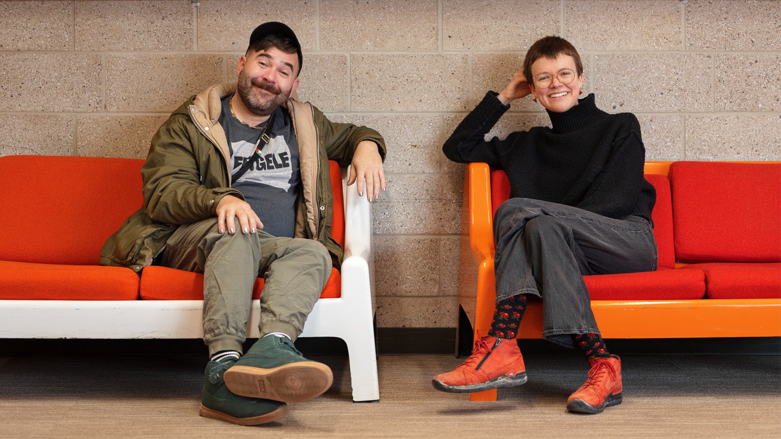 two smiling people sitting on couches with orange pillows