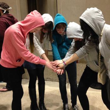 Five people in hooded sweatshirts form a circle and join their hands at the center
