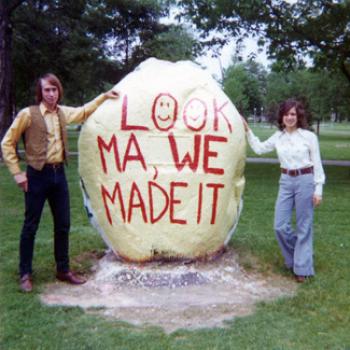 Two people in groovy 1970s attire pose by a boulder painted with the words, Look Ma, We Made It