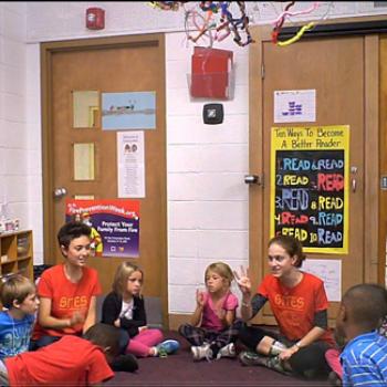 Two students wearing 'SITES' shirts sit in a circle with young children in a school classroom