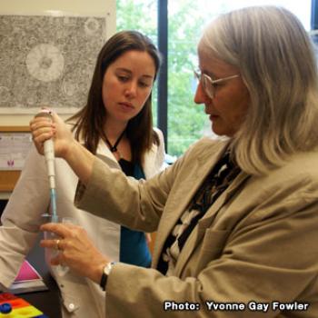 A professor handles a test tube while Olivia looks on.