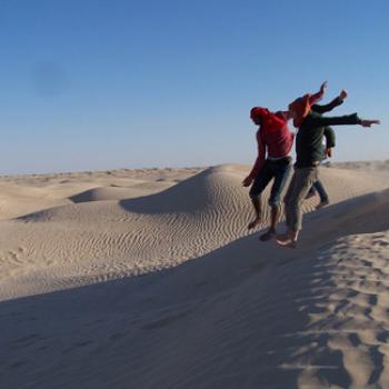 With only sand visible to the horizon, three people jump off a dune.