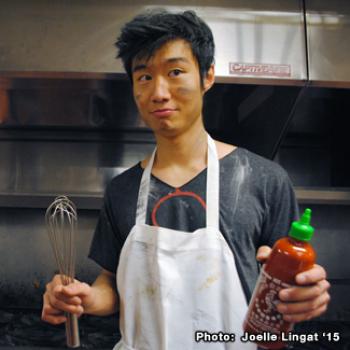 Wearing an apron, Weelic holds up a bottle of siracha and a whisk.