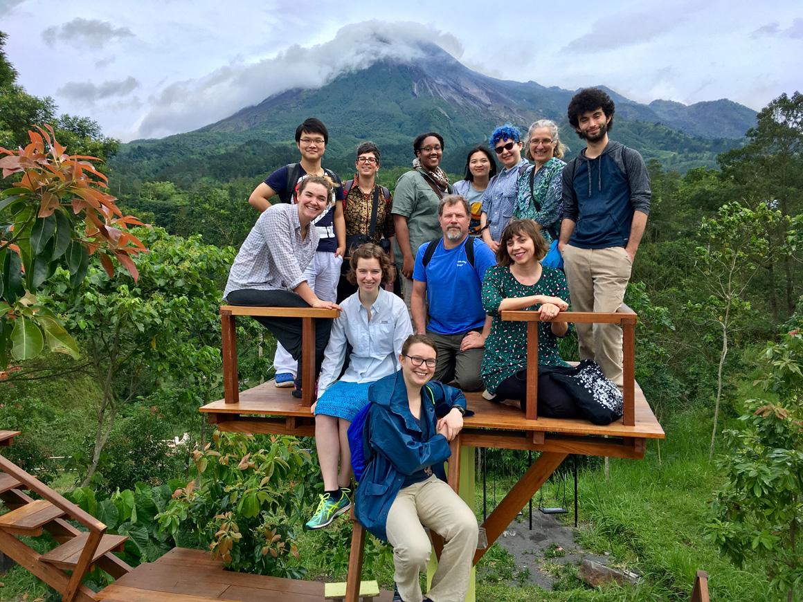 A dozen people pose on a wooden platform overlooking a lush forest. There is a mountain in the background.