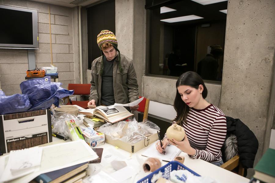At a table covered with papers, books, and boxes, two students examine an artifact.