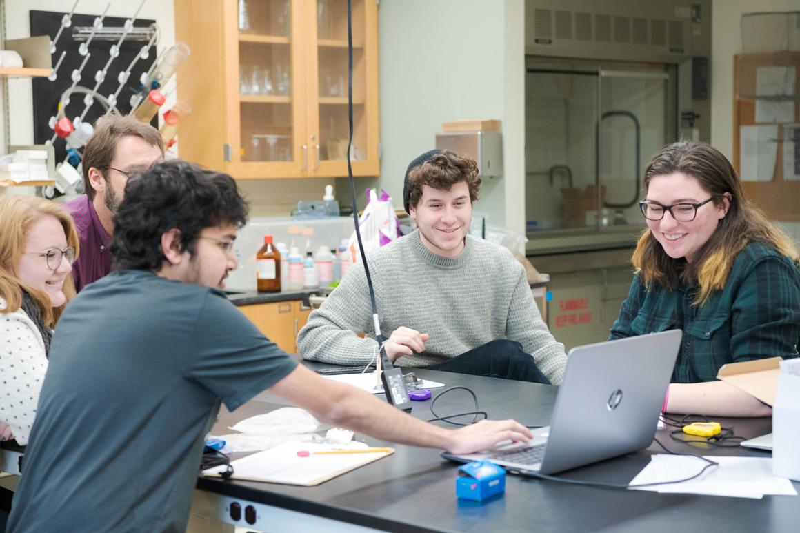Sitting around a lab bench, students and a professor look at a computer screen together.