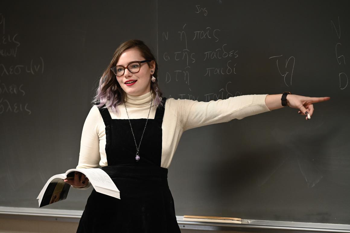 A student holds an open book while pointing something out on the blackboard.
