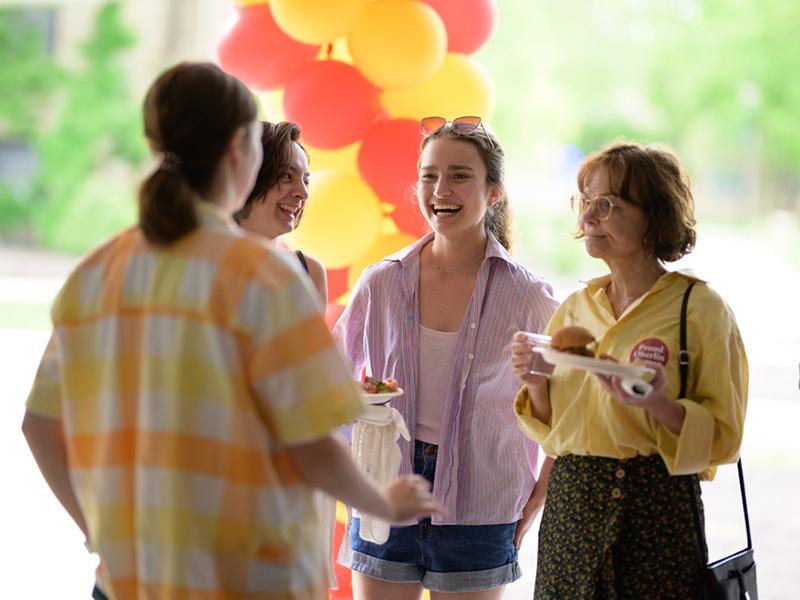 A small group, including a mom and daughter, talk and hold plates of food near a column of balloons. The mom is wearing a button that says 'Proud Oberlin Parent'.