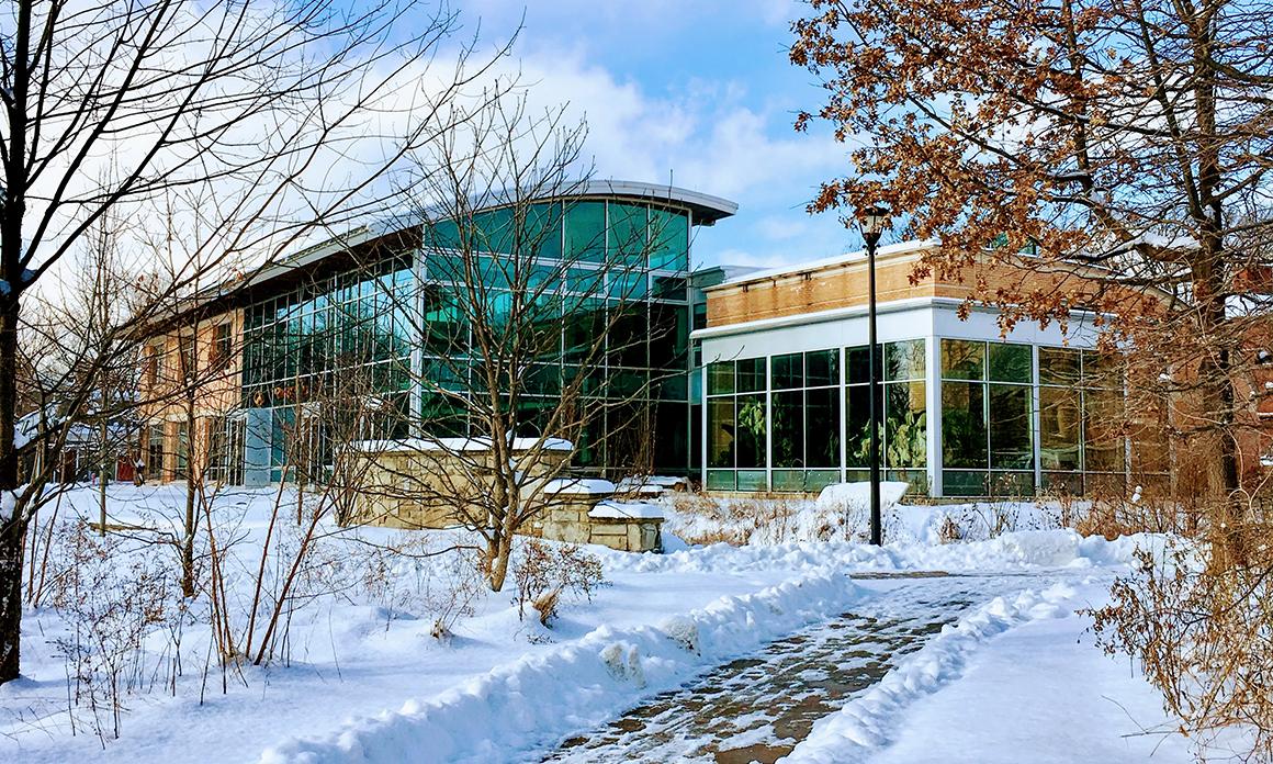 Exterior of a glass-sided, 2-story building on a sunny winter day. There is snow on the ground, and the path leading to the building has been shoveled.
