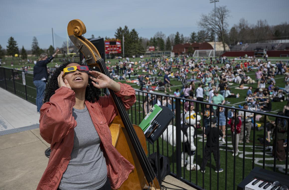 A young woman with a double bass looks at the solar eclipse through eclipse glasses, while a field of people behind her do the same.