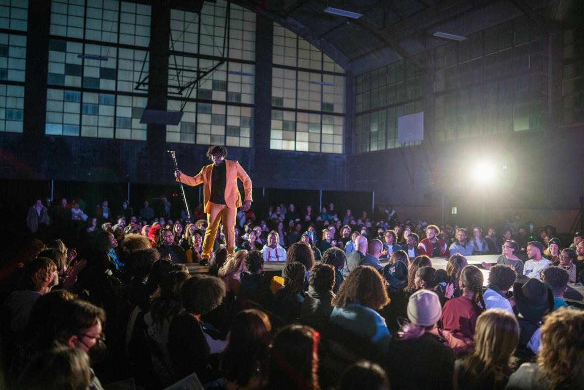 A male student poses in front of an audience for a fashion show in a gymnasium.