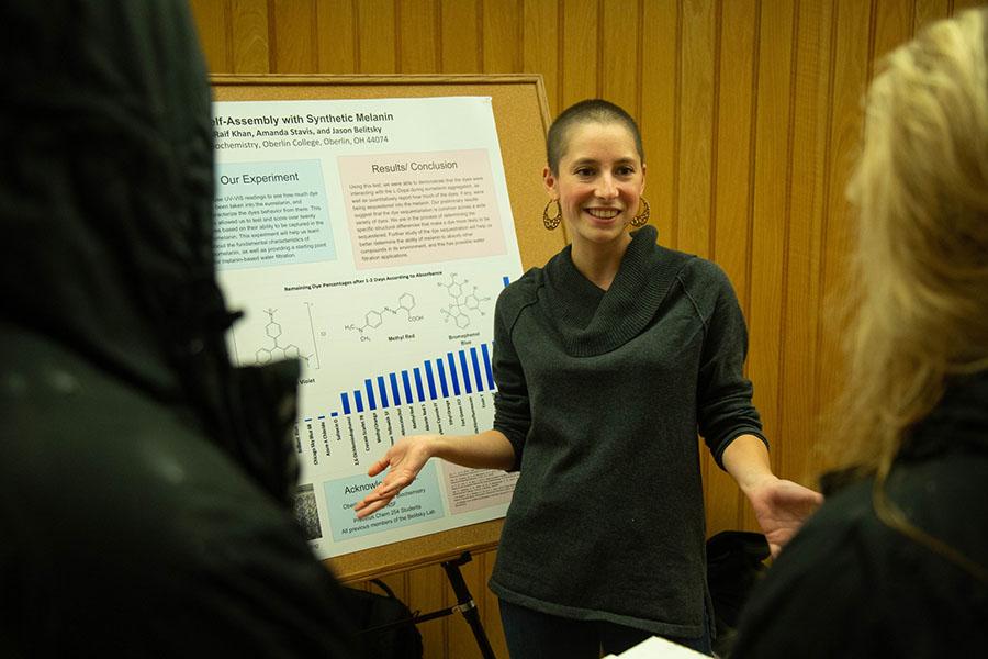 A student speaks to a small crowd in front of their poster presentation