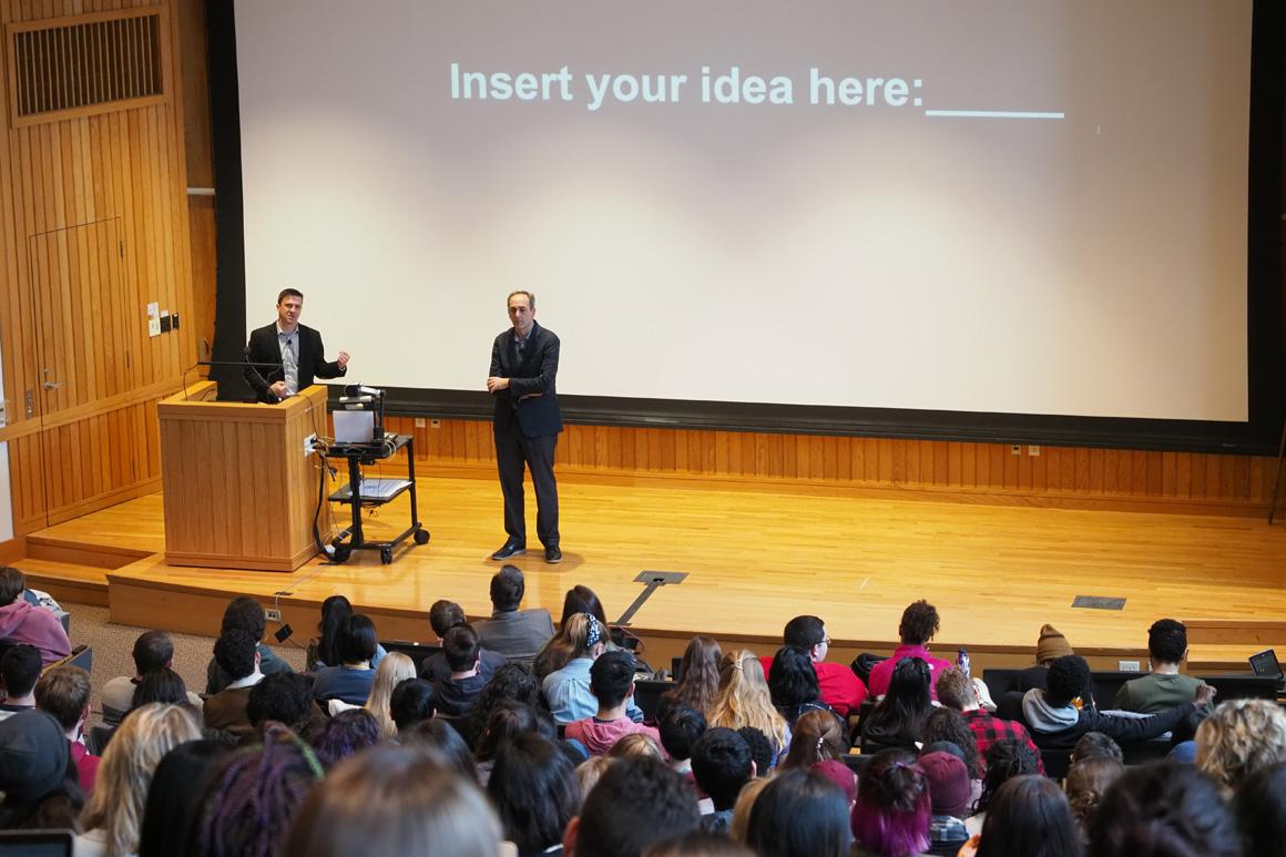 Two men present to a large class. The presentation slide reads, 'Insert your idea here' followed by a blank space.