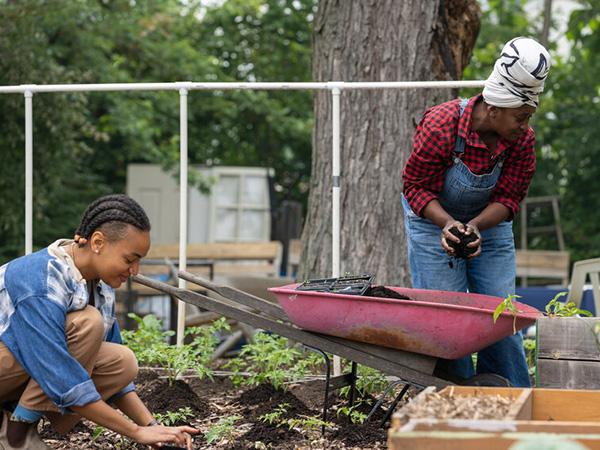 Two Black women, one older and one younger, work together tending to plants in a garden.