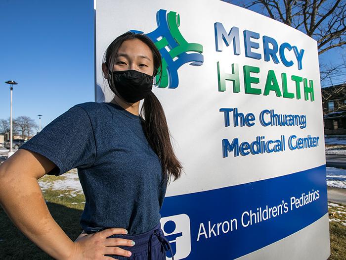 A student stands proudly next to a Mercy Health sign at Chuang Medical Center, Akron Children’s Pediatrics.