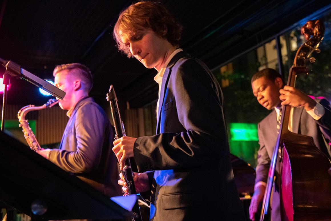 Three ensemble members—sax, clarinet, and bass—perform under colorful stage lights.