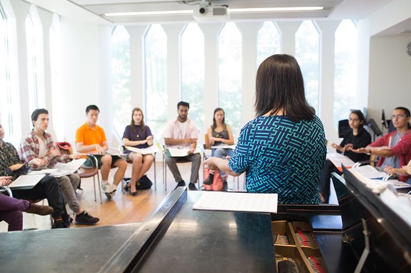 Students seated in a bright classroom where the professor is standing by a grand piano.