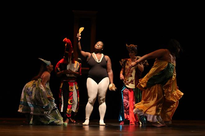 Dancers on stage. A woman gazes into a handheld mirror, surrounded by others in colorful costumes.