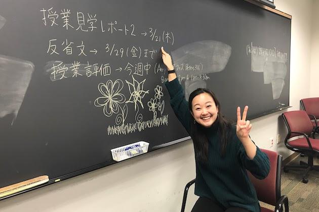 A lecturer of Japanese smiling in front of a blackboard.