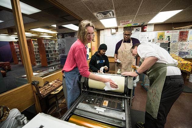 With library stacks in the background, 4 people wearing aprons set up a letterpress machine.