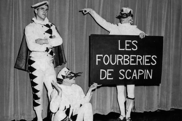 Three people on stage, one of them holding a sign that reads, "Les fourberies de scapin." Black and white.
