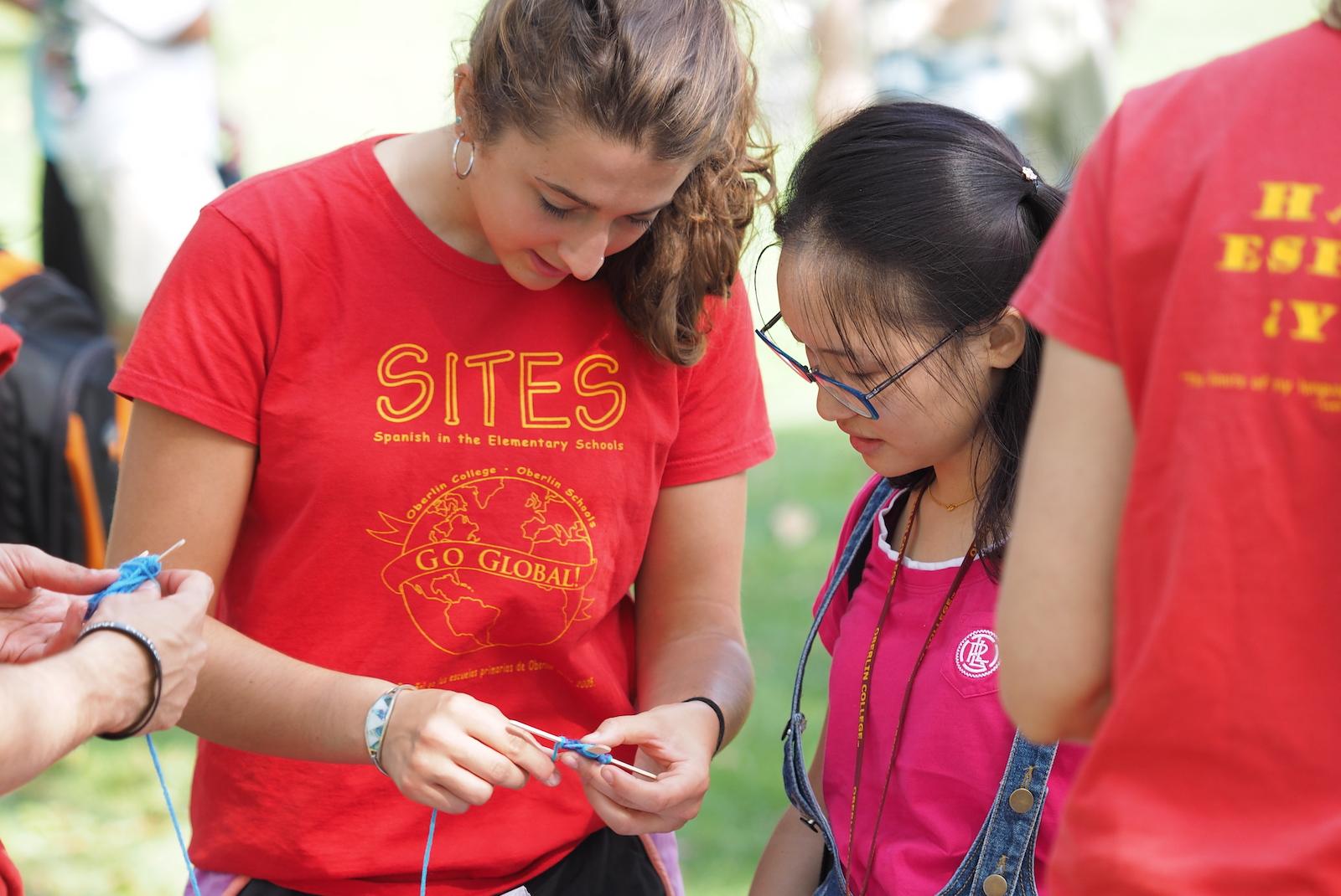 young woman shows student how to string a necklace.