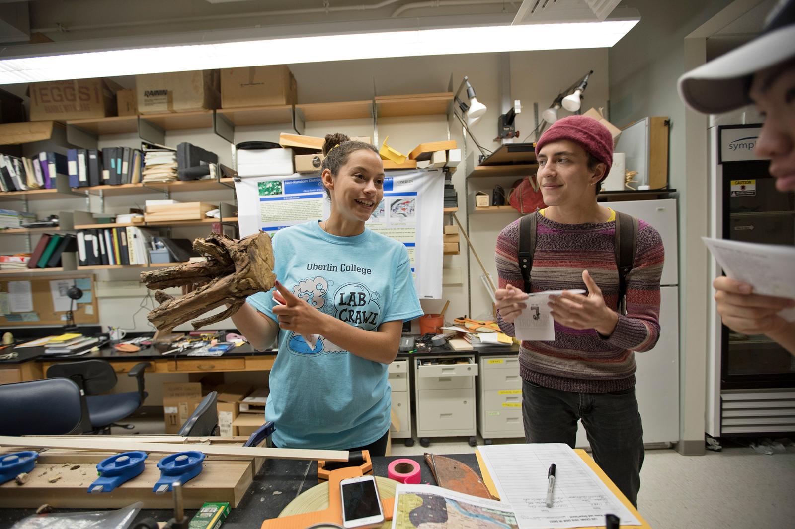 At a lab table, a student in a 'Lab Crawl' t-shirt holds up part of a tree branch or stump.