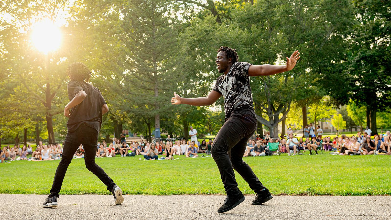 Two men in street clothes dance on the pavement in a park, in front of an audience seated in the grass.