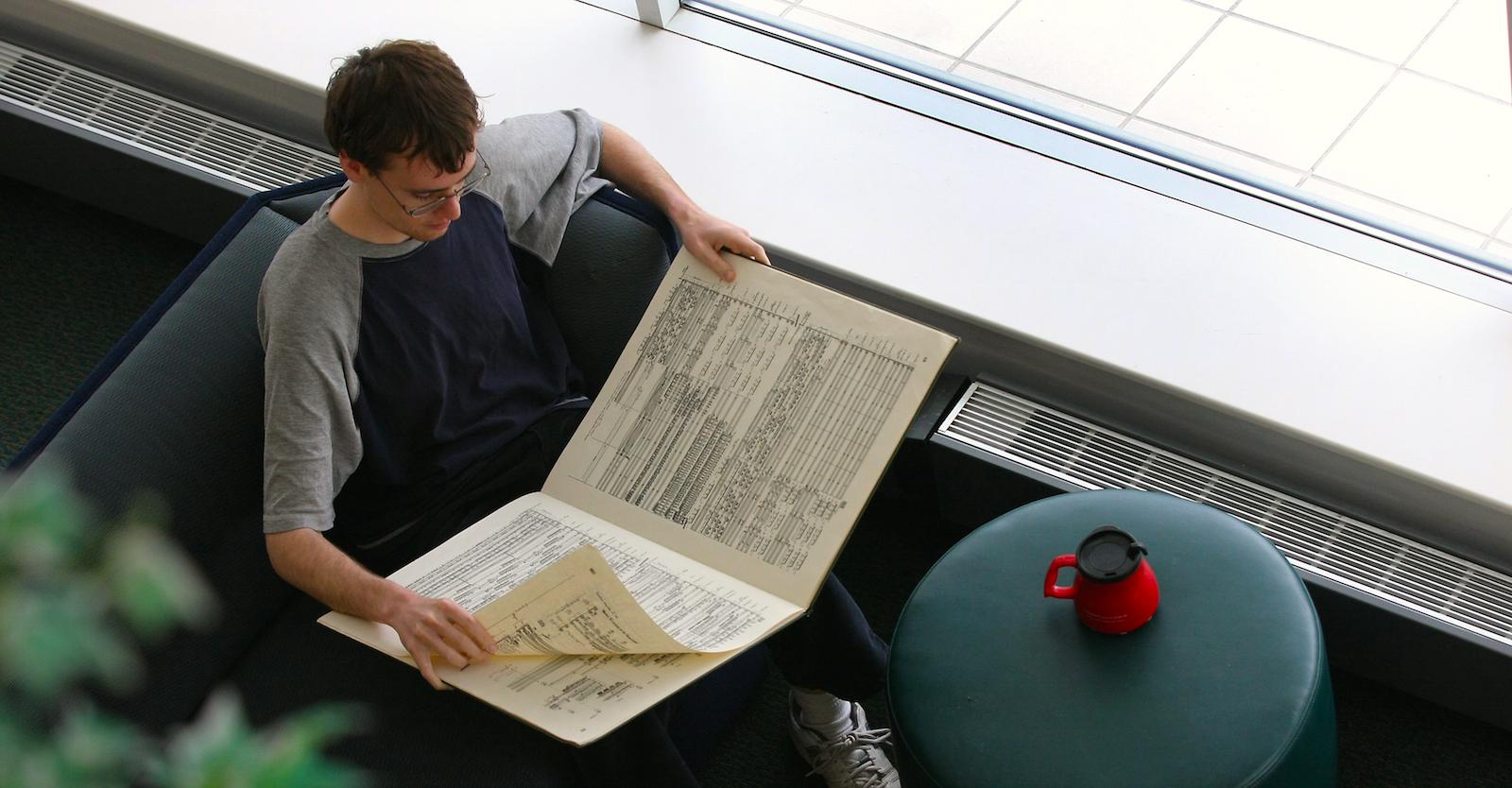 A student reviews a large book while comfortably seated. A travel mug sits on a small table.