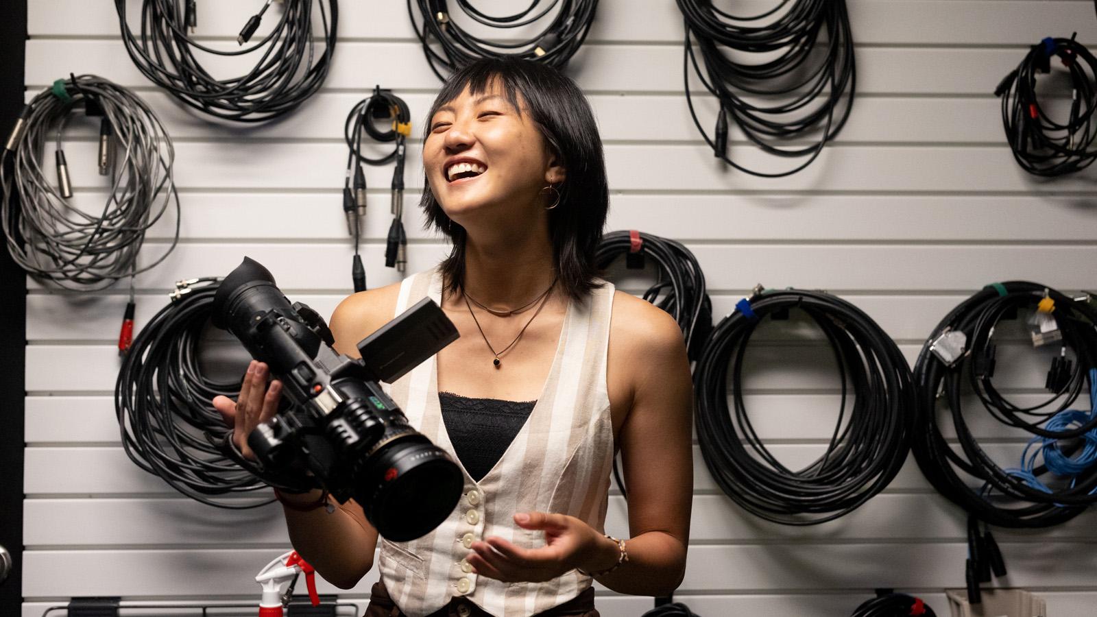 Standing in front of a wall where bundles of cables hang on hooks, a student holds a video camera and smiles.