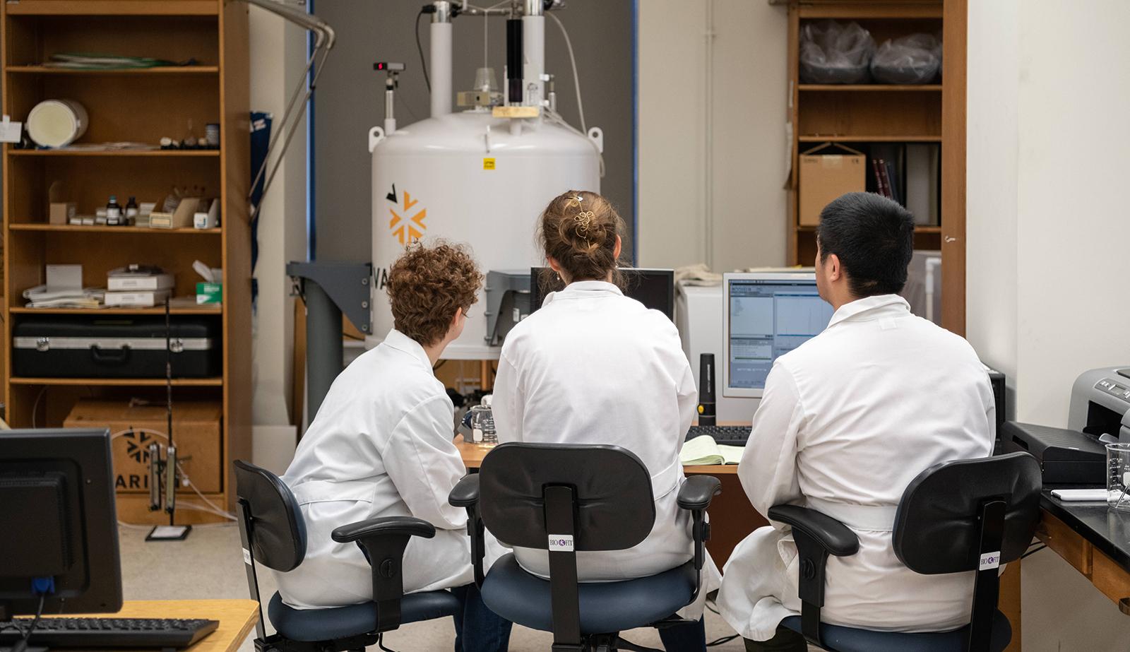 Three people in white lab coats sit in chairs facing away from the camera
