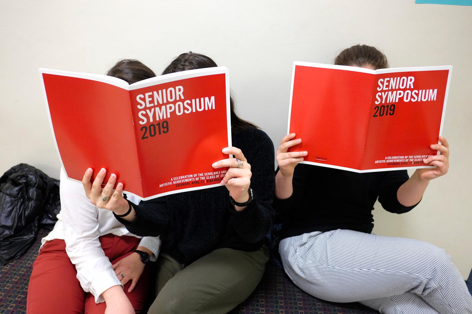 Three people read the 2019 research symposium program booklet, with the cover right in front of their faces.