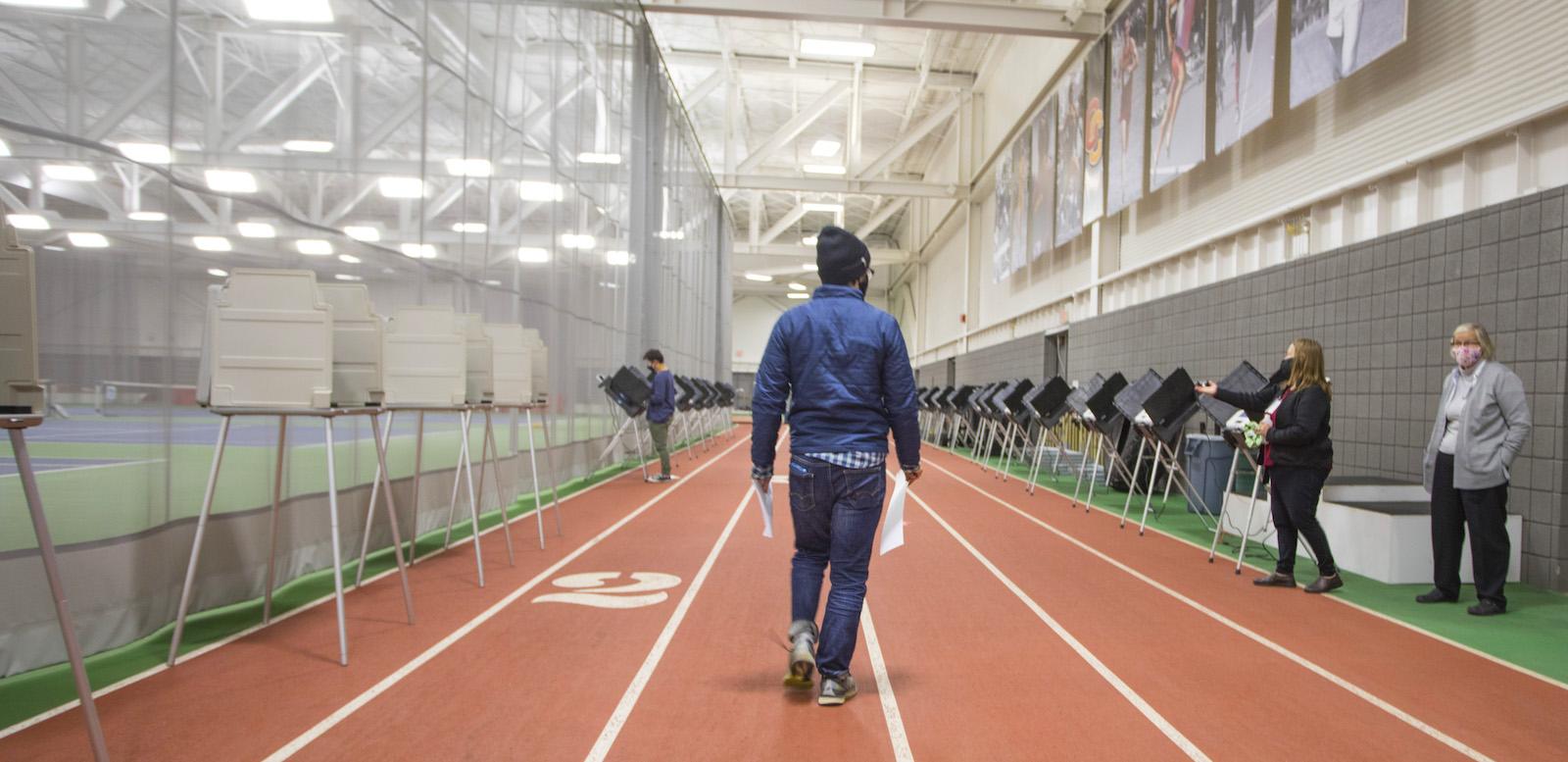 A student wearing a protective mask walks on an indoor track toward a voting booth.