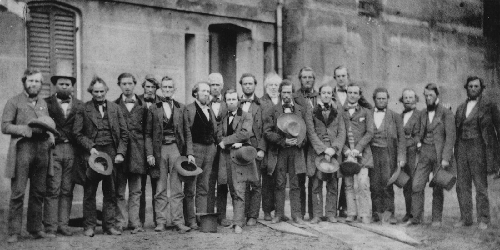 Historical photo of 20 men standing together, holding their hats.