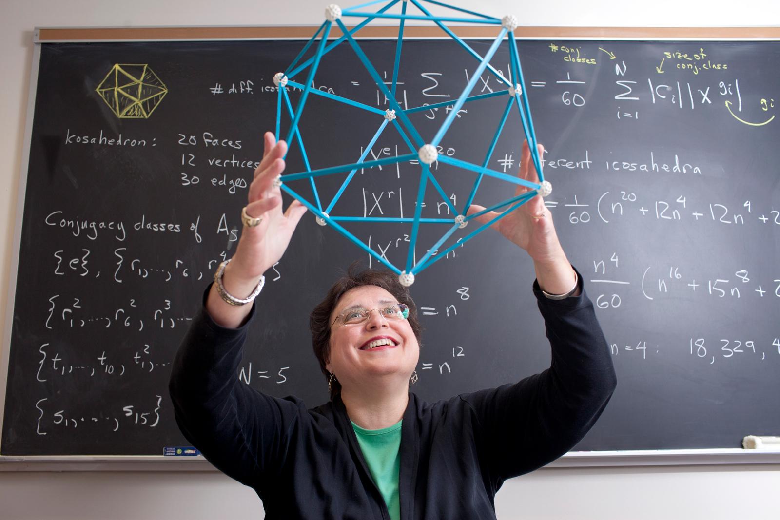 A professor holds a geometric model above her head. The blackboard behind her is covered in equations.