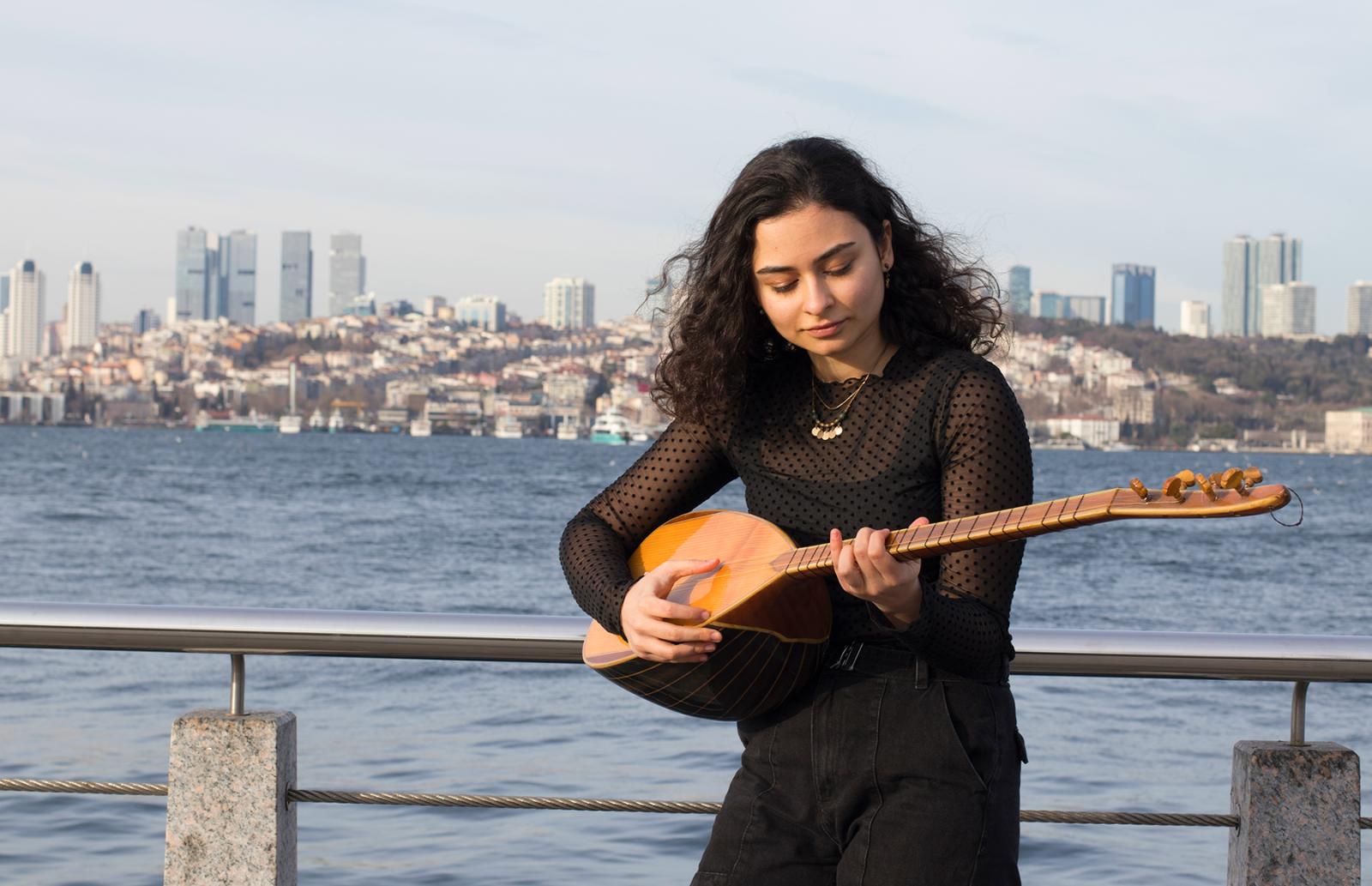 A woman plays a stringed instrument, with water and a city skyline behind her.