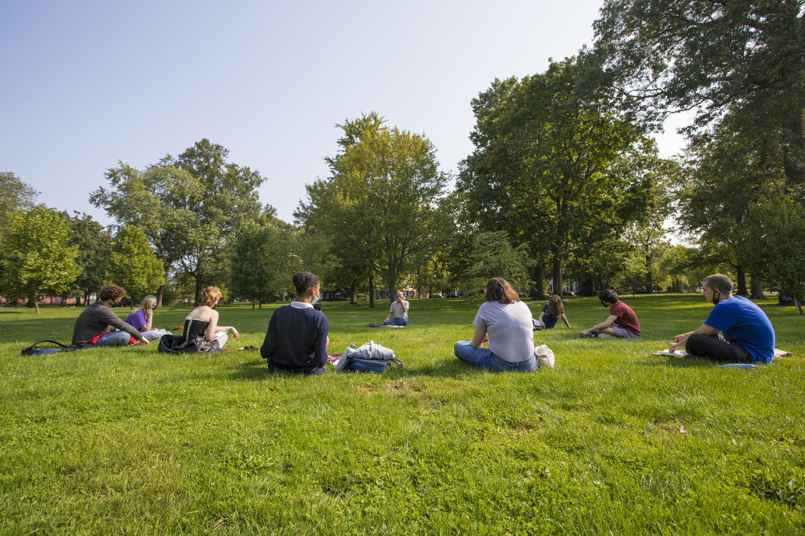 Students sitting in a circle in the grass, spaced widely apart from one another.