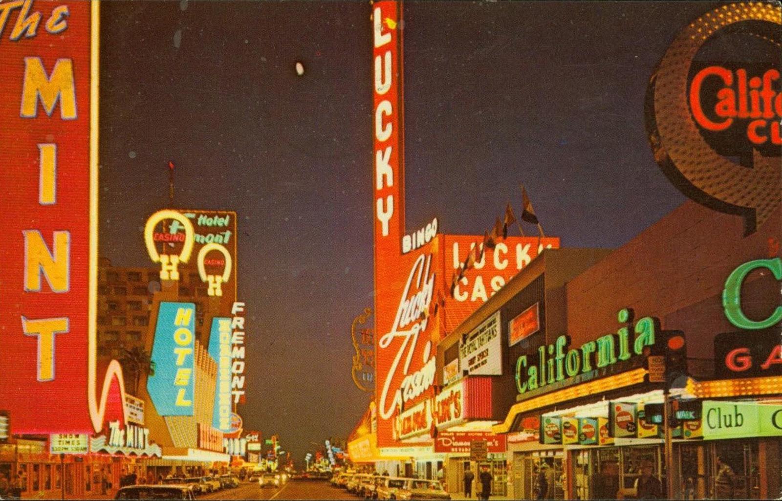 A 1950s city street with many lit up signs including Lucky Casino, The Mint, and hotels.