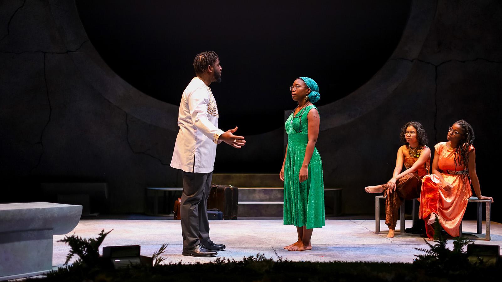 On stage, a man wearing black and white speaks to a woman in a green dress. Two women look on, wearing bronze and orange dresses.