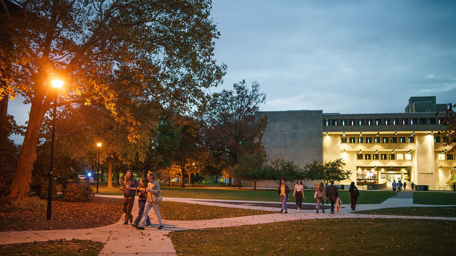 A campus quad at dusk. Students walk along a path in front of an illuminated building.