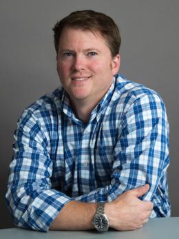 Man sitting against a desk in blue plaid shirt.