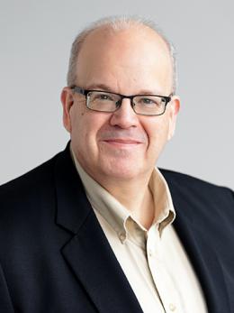 white man with silver thinning hair, wearing rectangular-framed glasses, an ecru open collared dress shirt, and black wool sport jacket