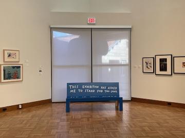Finnegan Shannon's "Do you want us here or not (MHR)—Bench," a blue bench with hand-painted text that reads, “This exhibition has asked me to stand for too long. Sit if you agree," is shown in a wideview of the gallery with multiple hung artworks.
